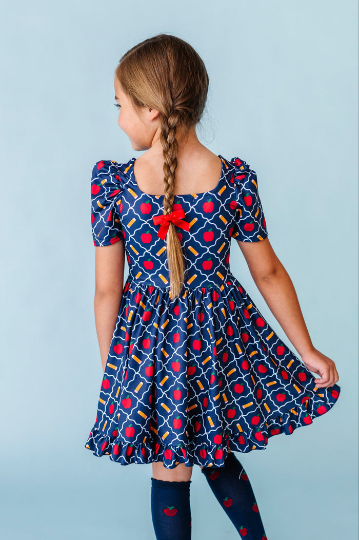 back view of little girl in braids wearing square neck navy school print twirl dress with matching red pigtail bows and apple knee socks