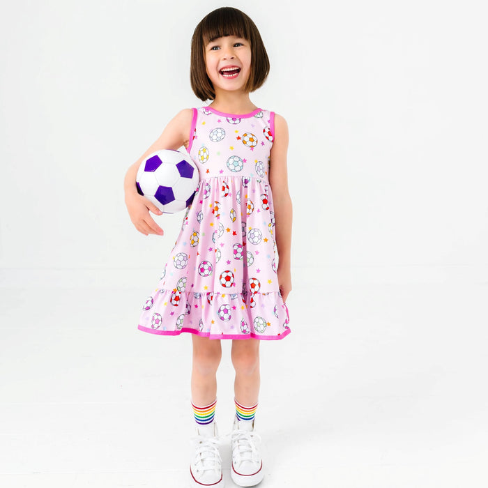 little girl holding soccer ball in pink sleeveless twirl dress with colorful soccer ball and star print paired with rainbow striped crew socks and white high tops