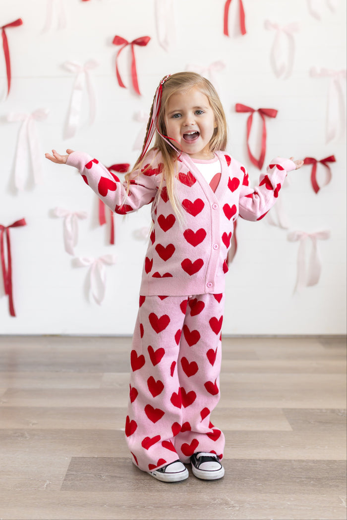 little girl in front of red and white bow covered wall with matching cotton knit cardigan and sweater pants featuring light pink background and red heart pattern