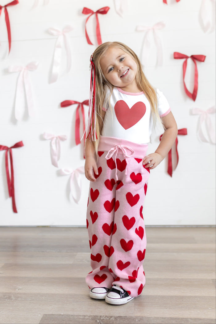 little girl in front of bow covered wall wearing white bamboo fitted top with red heart design paired with light pink cotton knit sweater pants with red hearts and matching ribbon hair clip