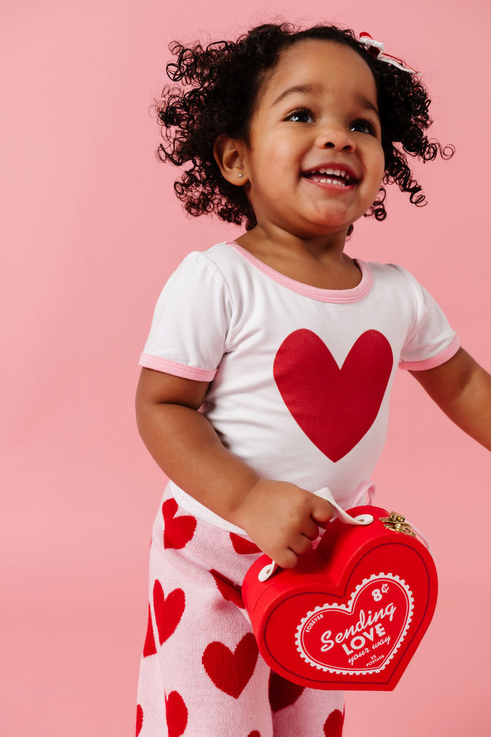 little girl holding heart shaped box in white bamboo top with red heart design paired with pink and red heart patterned sweater pants