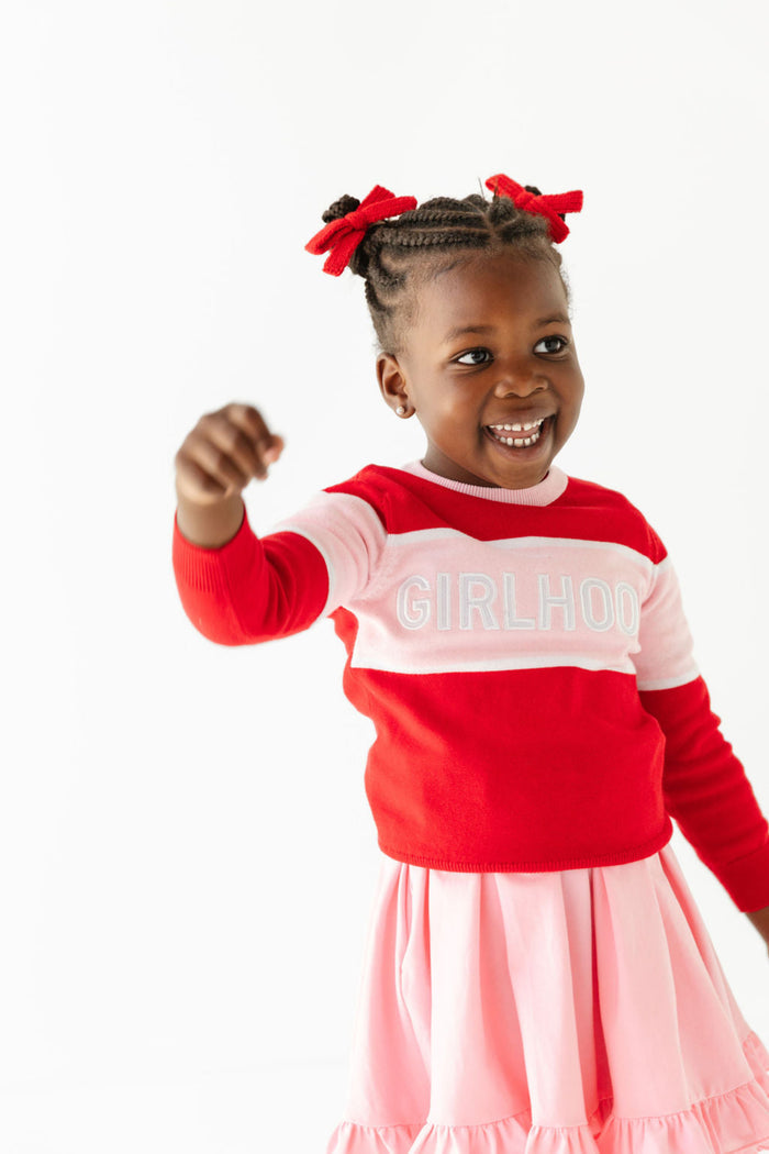 girl with pink twirl dress, red knit pigtail bows and coordinating red and pink GIRLHOOD design pullover sweater
