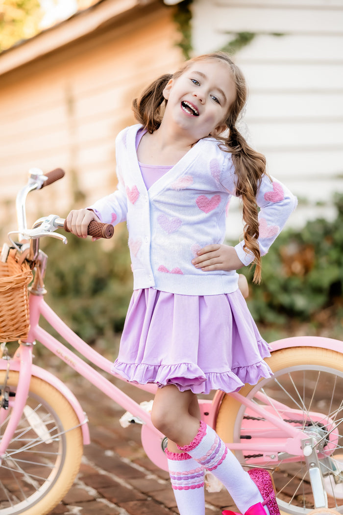 little girl with pink bicycle wearing lavender twirl dress layered with white cardigan with pastel fuzzy hearts and pastel bubble striped knee socks